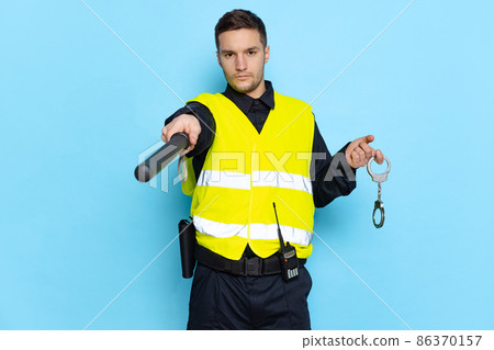 Half-length portrait of young male policeman officer wearing black uniform and warnvest posing isolated on blue background. Concept of job, caree, law and order. Half-length portrait of young male policeman officer wearing black uniform and warnvest posing isolated on blue background. Concept of job, caree, law and order. 86370157