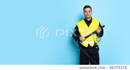 Half-length portrait of young male policeman officer wearing black uniform and warnvest posing isolated on blue background. Concept of job, caree, law and order. Half-length portrait of young male policeman officer wearing black uniform and warnvest posing isolated on blue background. Concept of job, caree, law and order. 86370158