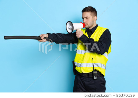 Portrait of young policeman officer wearing black uniform and warnvest shouting at megaphone isolated on blue background. Concept of job, caree, law and order. Portrait of young policeman officer wearing black uniform and warnvest shouting at megaphone isolated on blue background. Concept of job, caree, law and order. 86370159