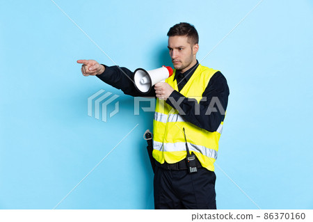 Portrait of young policeman officer wearing black uniform and warnvest shouting at megaphone isolated on blue background. Concept of job, caree, law and order. 86370160