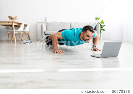 Smiling millennial european male doing push ups and watching online lesson on pc in minimalist living room 86370558