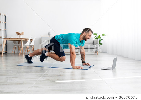 Glad smiling millennial european male in blue t-shirt doing leg exercises on mat with laptop watch video blog Glad smiling millennial european male in blue t-shirt doing leg exercises on mat with laptop watch video blog 86370573