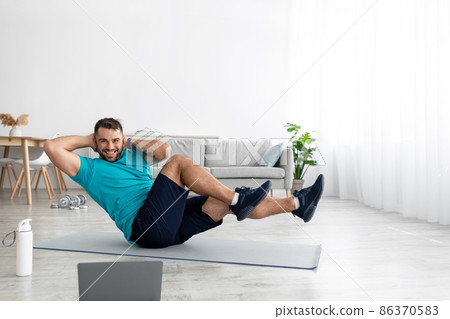 Cheerful young european male athlete in uniform doing abs exercises on mat with laptop in living room Cheerful young european male athlete in uniform doing abs exercises on mat with laptop in living room 86370583