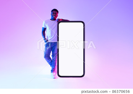 Smiling young black man standing near huge cellphone with empty white screen in neon light, mockup for mobile app Smiling young black man standing near huge cellphone with empty white screen in neon light, mockup for mobile app 86370656