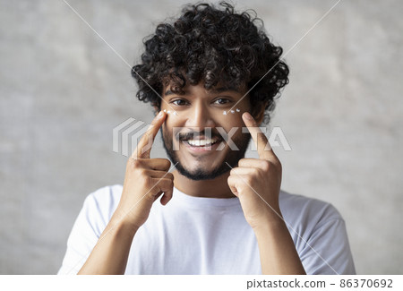 Portrait of happy indian man applying cream under eyes and smiling at camera, enjoying morning beauty routine 86370692