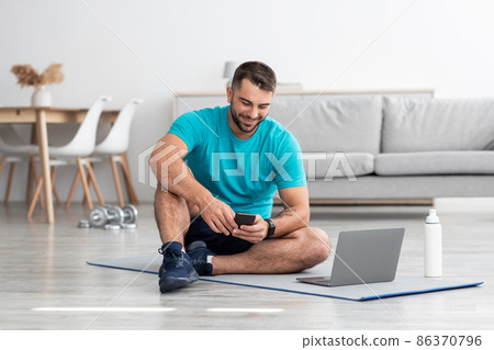 Cheerful millennial european man chatting on smartphone sits on mat in living room interior with bottle Cheerful millennial european man chatting on smartphone sits on mat in living room interior with bottle 86370796
