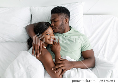 Cheerful young african american husband hugging wife and kissing on comfortable bed, resting, relax in bedroom Cheerful young african american husband hugging wife and kissing on comfortable bed, resting, relax in bedroom 86370864