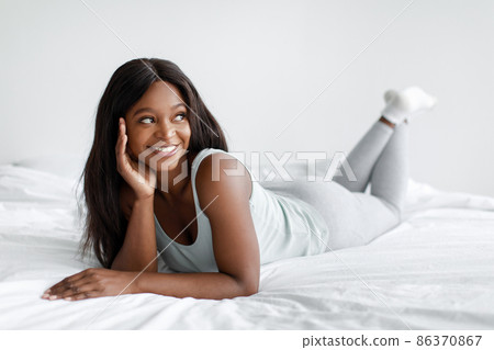 Smiling young african american woman in domestic clothes enjoy free time, lie on bed at weekend or vacation Smiling young african american woman in domestic clothes enjoy free time, lie on bed at weekend or vacation 86370867