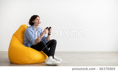 Young guy with joystick playing online video game, sitting in beanbag chair over light wall, panorama with free space Young guy with joystick playing online video game, sitting in beanbag chair over light wall, panorama with free space 86370869
