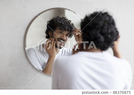 Male beauty and haircare routine. Handsome indian man touching beard and hair, looking in mirror in bathroom Male beauty and haircare routine. Handsome indian man touching beard and hair, looking in mirror in bathroom 86370875