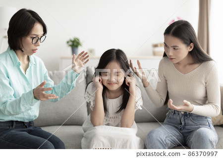 Asian girl covering ears, trying to hide from mother and grandmother scolding her at home 86370997