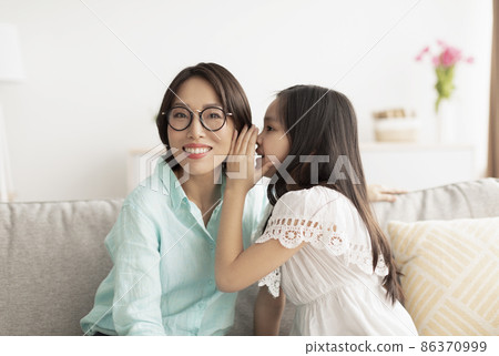 Cute little Asian girl whispering something into her grandmother's ear, sitting on couch at home Cute little Asian girl whispering something into her grandmother's ear, sitting on couch at home 86370999