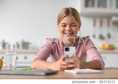 Cheerful caucasian teen girl student chatting at phone, sits at table in kitchen interior Cheerful caucasian teen girl student chatting at phone, sits at table in kitchen interior 86371013