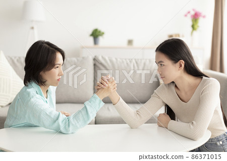 Mature Asian mother and adult daughter arm wrestling, looking each other in eyes, fighting at home Mature Asian mother and adult daughter arm wrestling, looking each other in eyes, fighting at home 86371015