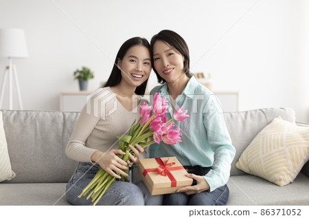Adult Asian daughter with her mature mother celebrating Women's Day, sitting on couch with tulips and gift at home 86371052