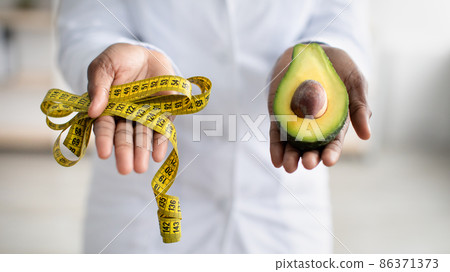 Healthy lifestyle concept. Black female dietitian holding half of avocado fruit and measure tape, closeup, panorama 86371373