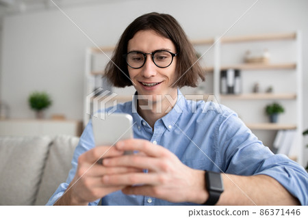 Positive young man sitting on couch with smartphone, chatting with ladies, using dating mobile application on cellphone Positive young man sitting on couch with smartphone, chatting with ladies, using dating mobile application on cellphone 86371446