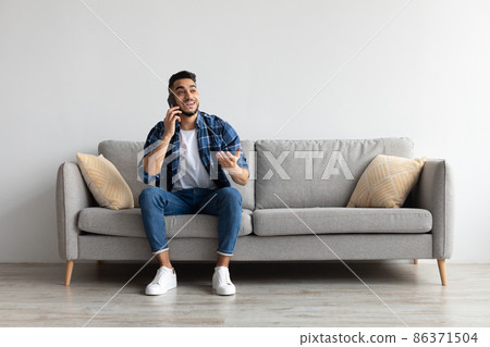 Cheerful Arab man talking on cellphone sitting on couch Cheerful Arab man talking on cellphone sitting on couch 86371504