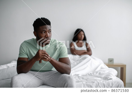 Depressed sad young black man ignores offended lady after quarrel sits on bed in bedroom interior, copy space Depressed sad young black man ignores offended lady after quarrel sits on bed in bedroom interior, copy space 86371580