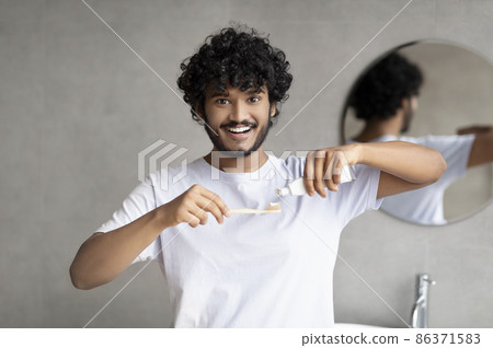 Teeth cleaning concept. Young indian man applying toothpaste on toothbrush, brushing teeth in bathroom in the morning Teeth cleaning concept. Young indian man applying toothpaste on toothbrush, brushing teeth in bathroom in the morning 86371583