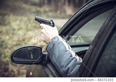 A man with a gun driving a car, a man's hand with a gun in his hand leaned out of the car window 86372103