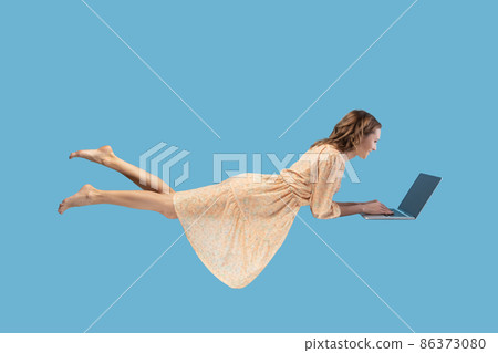 Hovering in air. Happy cheerful girl in yellow dress levitating with laptop, typing keyboard, reading good news message on computer while flying in mid-air. studio shot isolated on blue background 86373080