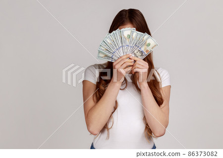 Unknown woman hiding face behind bunch of dollar banknotes, anonymous person holding money, lottery win, big profit, wearing white T-shirt. Indoor studio shot isolated on gray background. 86373082
