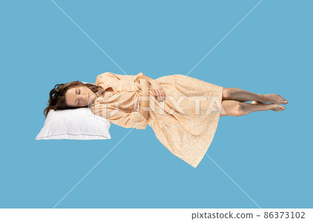 Sleeping beauty hovering in air. Relaxed girl in yellow dress lying comfortably on pillow levitating, keeping eyes closed, watching dreams. full length studio shot isolated on blue background, indoor 86373102