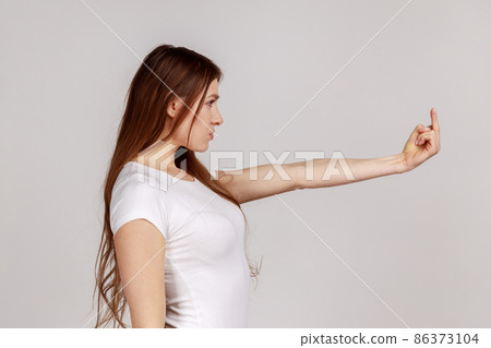 Side view of annoyed woman showing middle finger to camera, expressing protest hate and disrespect, gesturing rude symbol, wearing white T-shirt. Indoor studio shot isolated on gray background. 86373104