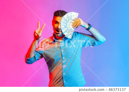 Portrait of rich funny man in shirt covering half of face with dollars, looking at camera showing vi sign, being happy to win lottery. Indoor studio shot isolated on colorful neon light background. 86373137