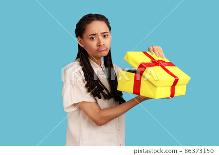 Portrait of unhappy upset woman with black dreadlocks looking into gift box, opening present and looking at camera with sorrow, wearing white shirt. Indoor studio shot isolated on blue background. 86373150