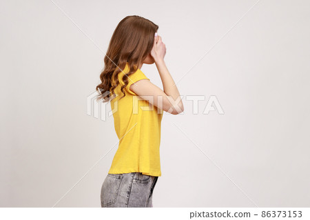 I'm not watching. Profile portrait of slim girl in yellow casual T- shirt covering eyes with hand, frightened to look, sees disgusting things. Indoor studio shot isolated on gray background. 86373153