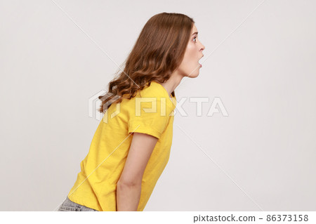 Side view of shocked beautiful young girl with brown wavy hair in yellow T-shirt standing and with surprised face, hearing shocking news. Indoor studio shot isolated on gray background. Side view of shocked beautiful young girl with brown wavy hair in yellow T-shirt standing and with surprised face, hearing shocking news. Indoor studio shot isolated on gray background. 86373158