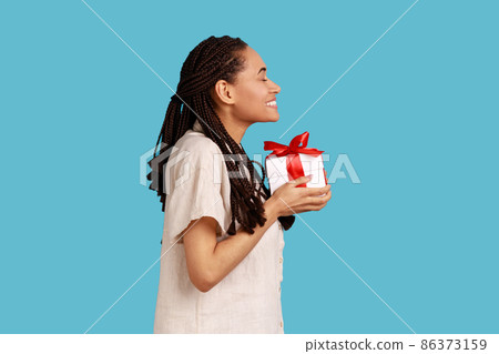 Side view of cute charming woman with black dreadlocks holding present box, congratulating, extremely happy to get gift, wearing white shirt. Indoor studio shot isolated on blue background. Side view of cute charming woman with black dreadlocks holding present box, congratulating, extremely happy to get gift, wearing white shirt. Indoor studio shot isolated on blue background. 86373159