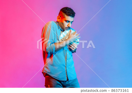 Man in shirt embracing dollar banknotes looking at camera with greedy expression, afraid to lose money, obsessed with finances. Indoor studio shot isolated on colorful neon light background. 86373161