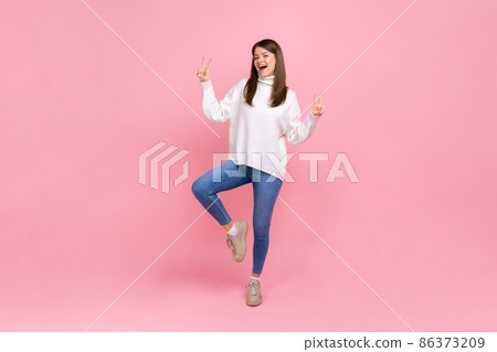 Full length portrait of happy woman doing victory gesture, showing peace, v sign with double fingers, wearing white casual style sweater. Indoor studio shot isolated on pink background. 86373209