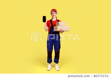 Full length portrait of positive delivery woman standing with cardboard packages and showing phone with blank screen for advertisement. Indoor studio shot isolated on yellow background. Full length portrait of positive delivery woman standing with cardboard packages and showing phone with blank screen for advertisement. Indoor studio shot isolated on yellow background. 86373210