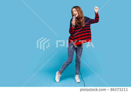 Full length portrait of woman wearing striped casual style sweater, singing in microphone and dancing, having fun in karaoke, performing alone. Indoor studio shot isolated on blue background. Full length portrait of woman wearing striped casual style sweater, singing in microphone and dancing, having fun in karaoke, performing alone. Indoor studio shot isolated on blue background. 86373211