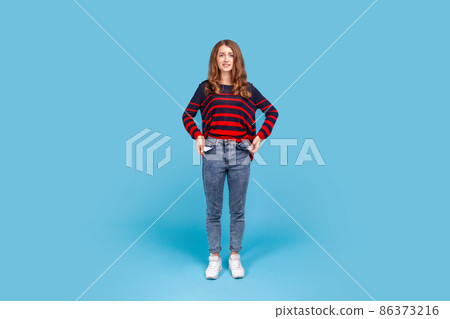 Full length portrait of poor woman, turning out empty pockets, worried about debts, no cash for living, wearing striped casual style sweater. Indoor studio shot isolated on blue background. Full length portrait of poor woman, turning out empty pockets, worried about debts, no cash for living, wearing striped casual style sweater. Indoor studio shot isolated on blue background. 86373216