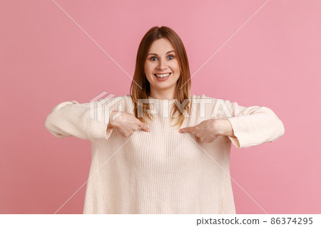 Portrait of blond woman pointing herself and looking at camera with toothy smile, proud of own success, achievement, wearing white sweater. Indoor studio shot isolated on pink background. 86374295