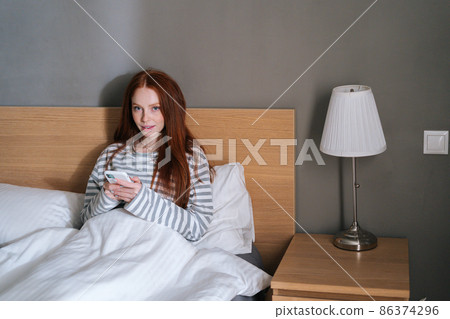 Front view of atractive young woman using mobile phone, typing online message on social media, looking at camera, sitting lying on cozy bed at evening in bedroom. 86374296
