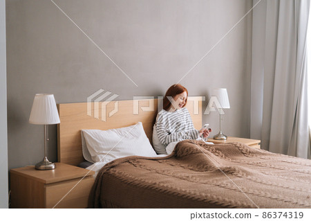 Remote view of happy young woman enjoy shopping online with credit card and mobile phone lying in bed at home. Smiling female holding credit card to make online payment at bedroom. Remote view of happy young woman enjoy shopping online with credit card and mobile phone lying in bed at home. Smiling female holding credit card to make online payment at bedroom. 86374319
