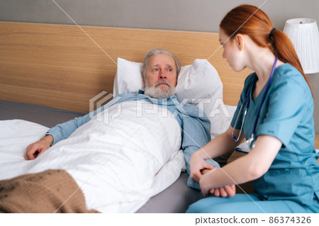 Female doctor measuring pulse on wrist to sick senior male patient lying on bed in hospital room. Nurse checking mature patient pulse at home. Elderly man getting medical consultation in hospice. 86374326