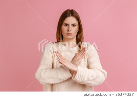 Portrait of blond woman standing with crossed hands, gesturing stop, finish, showing symbol of rejection, body language, wearing white sweater. Indoor studio shot isolated on pink background. 86374327
