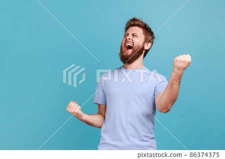 Portrait of overjoyed bearded man standing with excited expression, raising fists, screaming, shouting yeah, celebrating his victory, success. Indoor studio shot isolated on blue background. 86374375