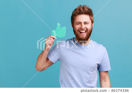 Portrait of extremely happy positive bearded man holding and showing like or thumbs up paper shape sign, looking at camera with toothy smile. Indoor studio shot isolated on blue background. 86374386