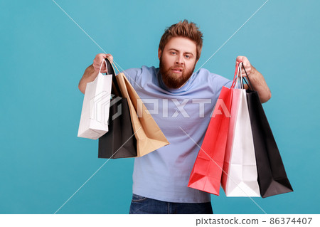 Portrait of tiered exhausted handsome bearded man raised arms with shopping bags,looking at camera, feels bored after going in mall. Indoor studio shot isolated on blue background. 86374407
