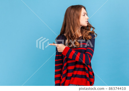 Get out. Portrait of angry resentful woman wearing striped casual style sweater, pointing way out and grimacing madly, ordering to leave, showing exit. Indoor studio shot isolated on blue background. 86374410