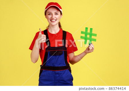 Portrait of worker woman standing holding green hashtag in hands, looking at camera and showing thumb up, wearing overalls and red cap. Indoor studio shot isolated on yellow background. Portrait of worker woman standing holding green hashtag in hands, looking at camera and showing thumb up, wearing overalls and red cap. Indoor studio shot isolated on yellow background. 86374414