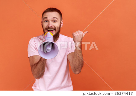 Portrait of bearded man holding megaphone and pointing aside, showing copyspace for advertisement with happy expression, wearing pink T-shirt. Indoor studio shot isolated on orange background. Portrait of bearded man holding megaphone and pointing aside, showing copyspace for advertisement with happy expression, wearing pink T-shirt. Indoor studio shot isolated on orange background. 86374417